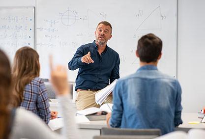 A man sits off the edge of a desk, talking to engaged students in front of him.